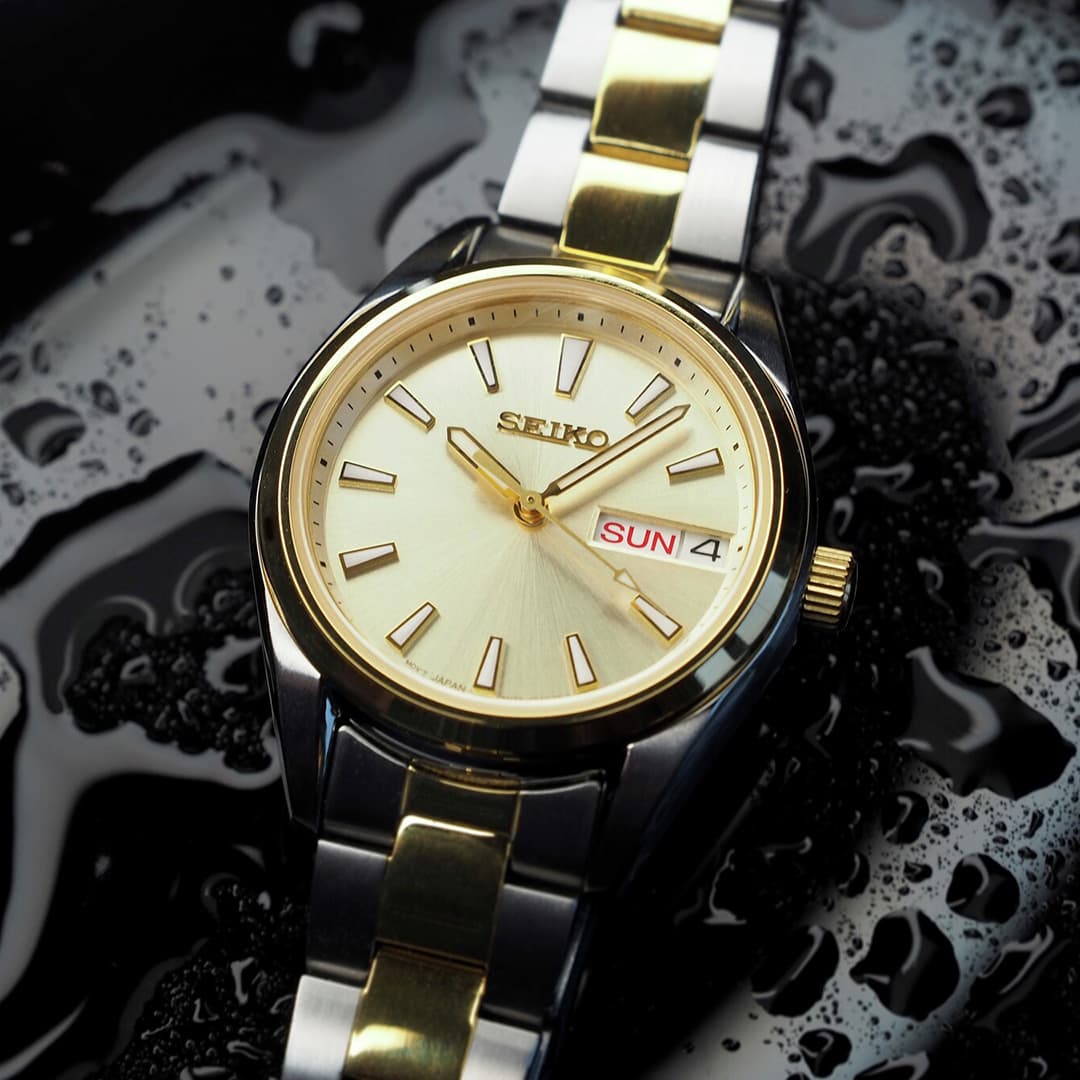 Et Seiko-armbåndsur i sølv og gull som viser dag og dato, mot en teksturert svart overflate med vanndråper.