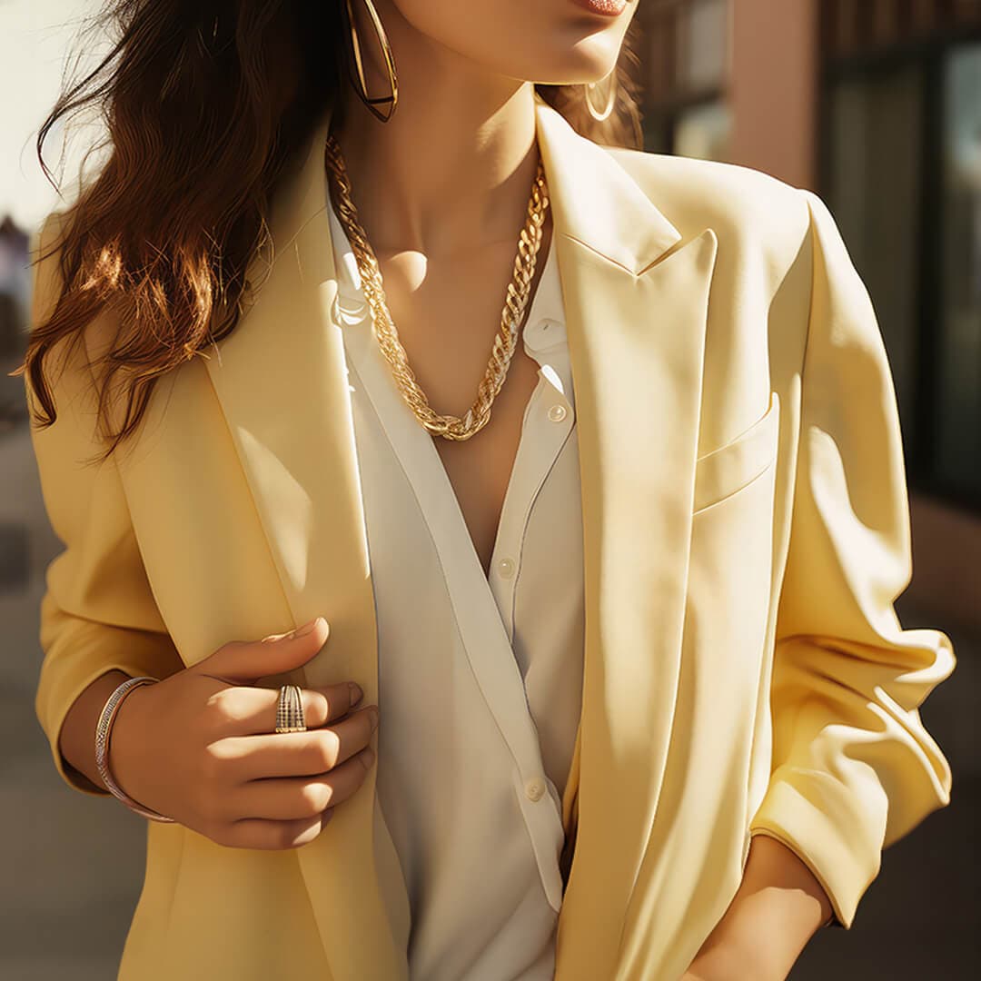 Kvinne i gul blazer og hvit skjorte, tilbehørt med store gulløredobber, halskjede og armbånd, stående utendørs i sollys.