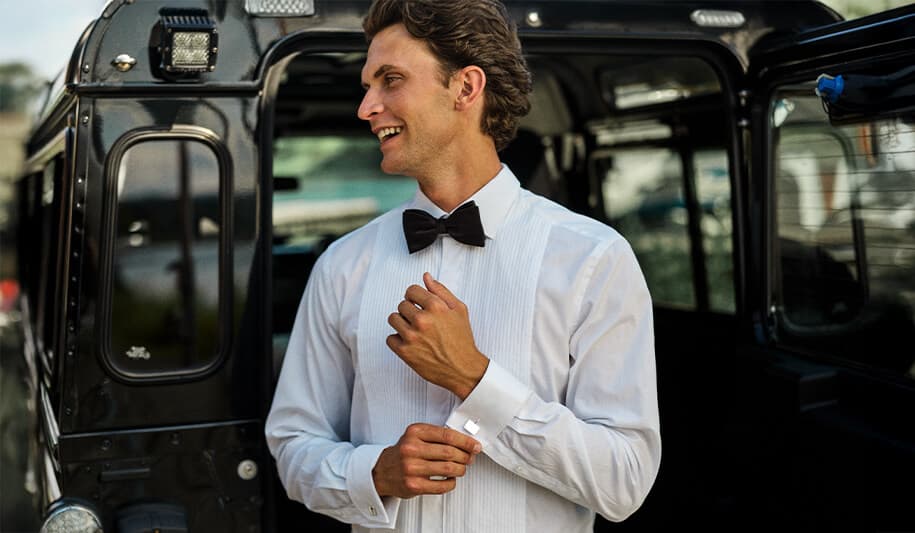 Mann i hvit penskjorte og sort sløyfe som justerer mansjettknappen sin, smilende, stående ved siden av et mørkt kjøretøy.