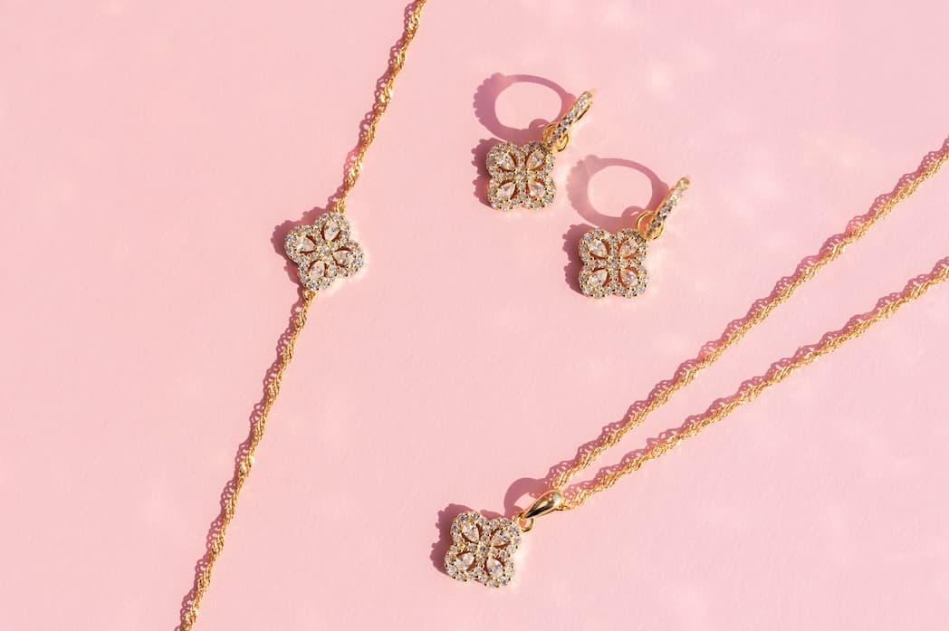 Gullsmykkesett med blomsterdiamantmønstre, inkludert et halskjede, armbånd og øredobber, vist på en rosa bakgrunn.