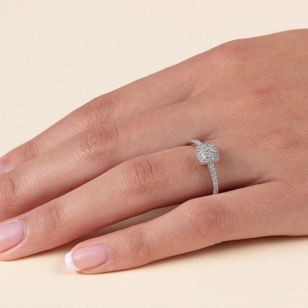 En hånd med en sølvring med firkantet diamantfatning, som hviler på en lys beige overflate.