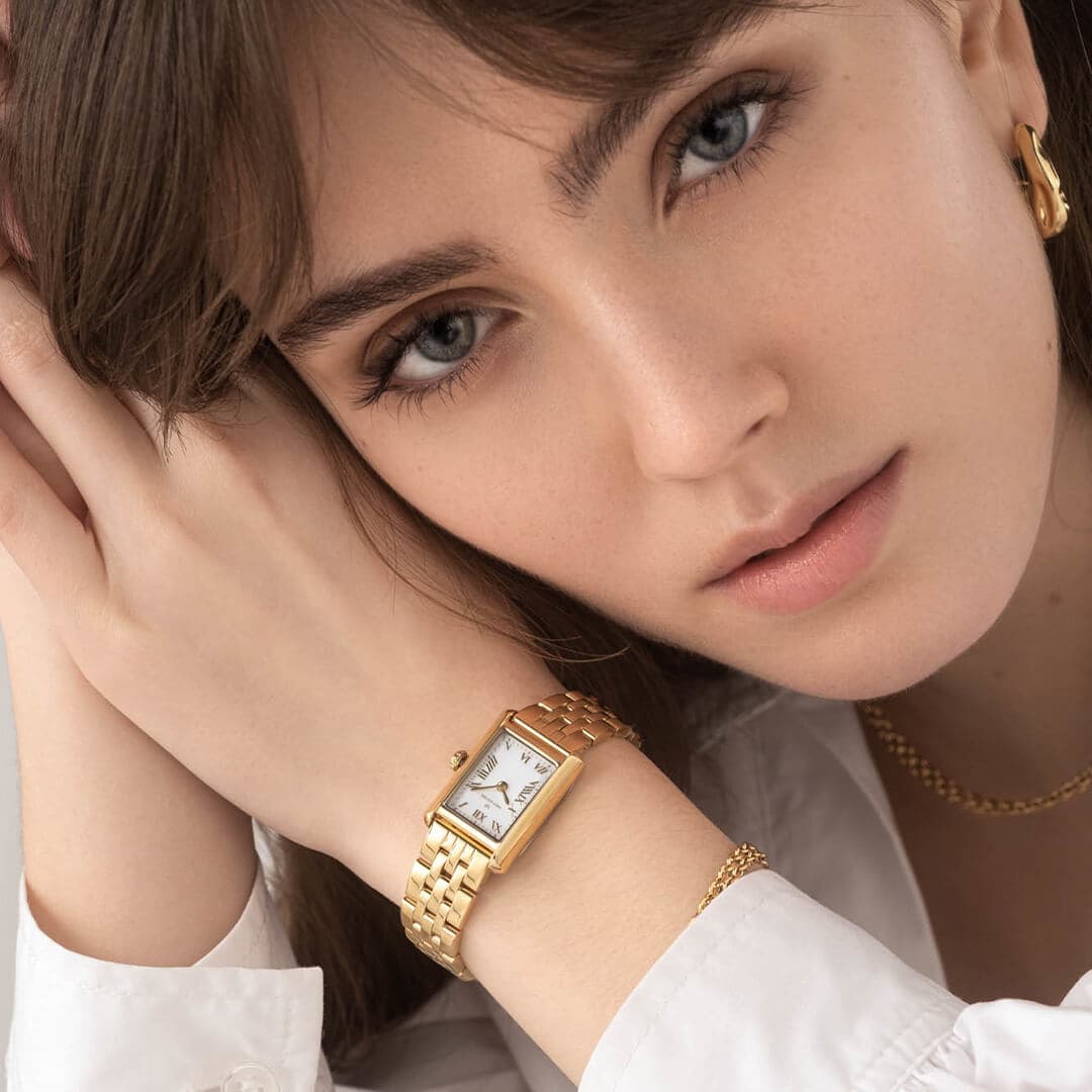 Kvinne med gullklokke, gullringeøreringer og et halskjede, hviler haken på hendene og ser inn i kameraet.