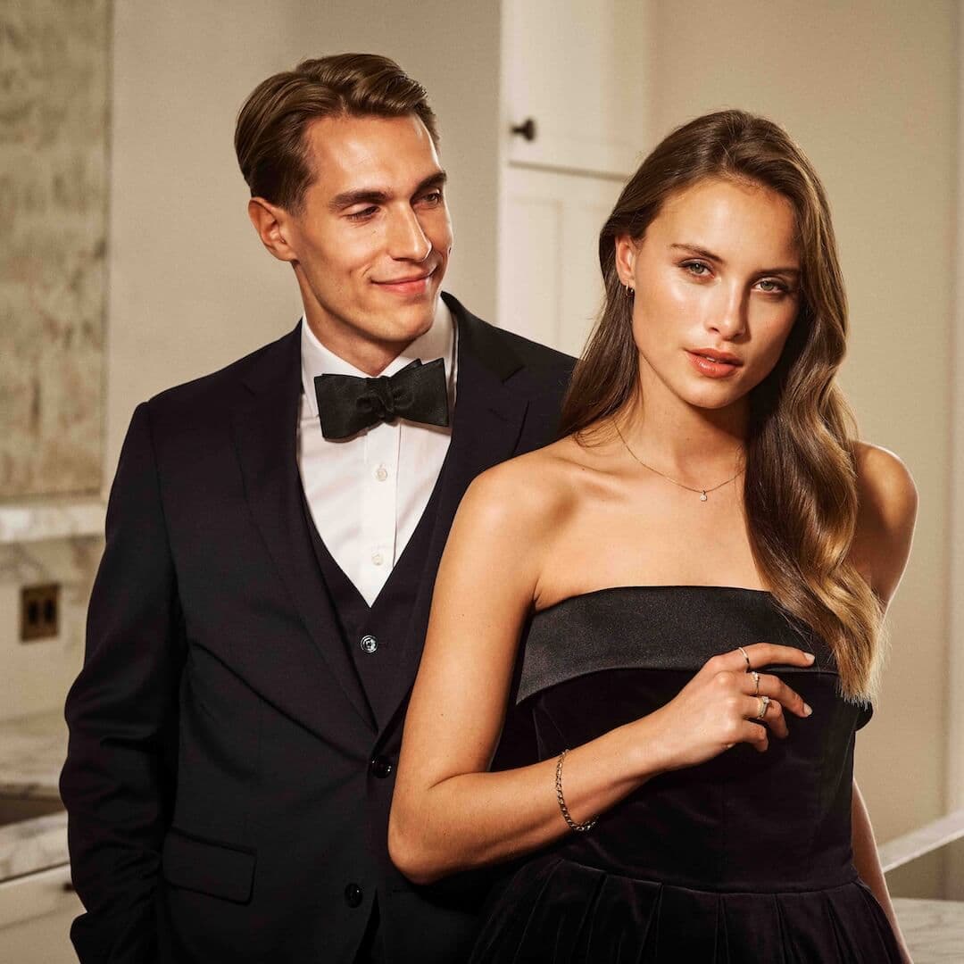 En mann i smoking smiler og står ved siden av en kvinne i en stroppeløs svart kjole, mens de poserer elegant i en innendørs setting.