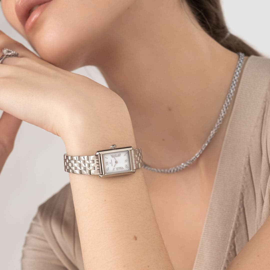 Kvinne med en sølvklokke med rektangulær urskive og et matchende halskjede, kledd i en beige topp.