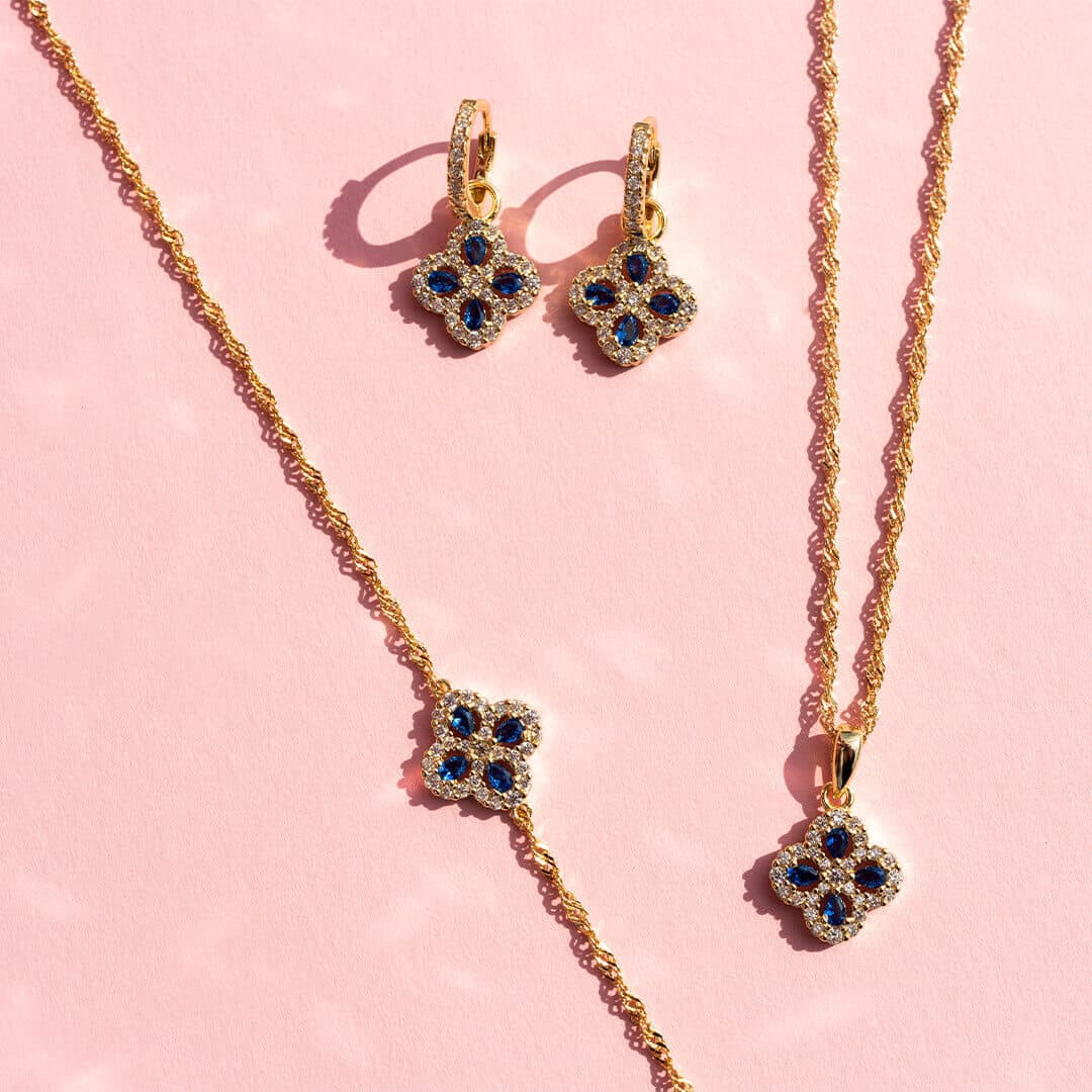 Gullsmykkesett med safiraksenterte blomsterdesign, inkludert et halskjede, armbånd og øredobber, vist på en rosa bakgrunn.