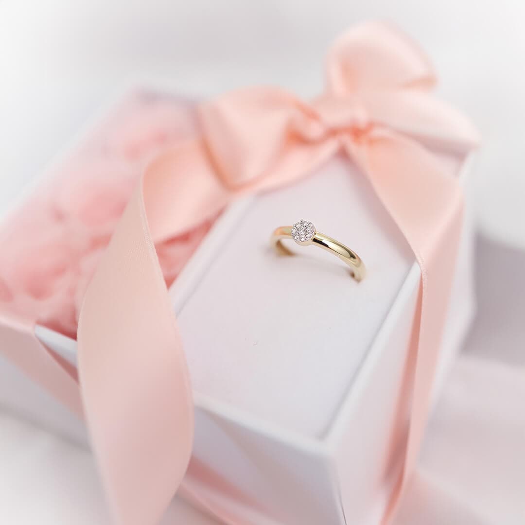 Gullring med en liten rund diamant på en hvit eske toppet med et rosa bånd, omgitt av rosa roseblader.