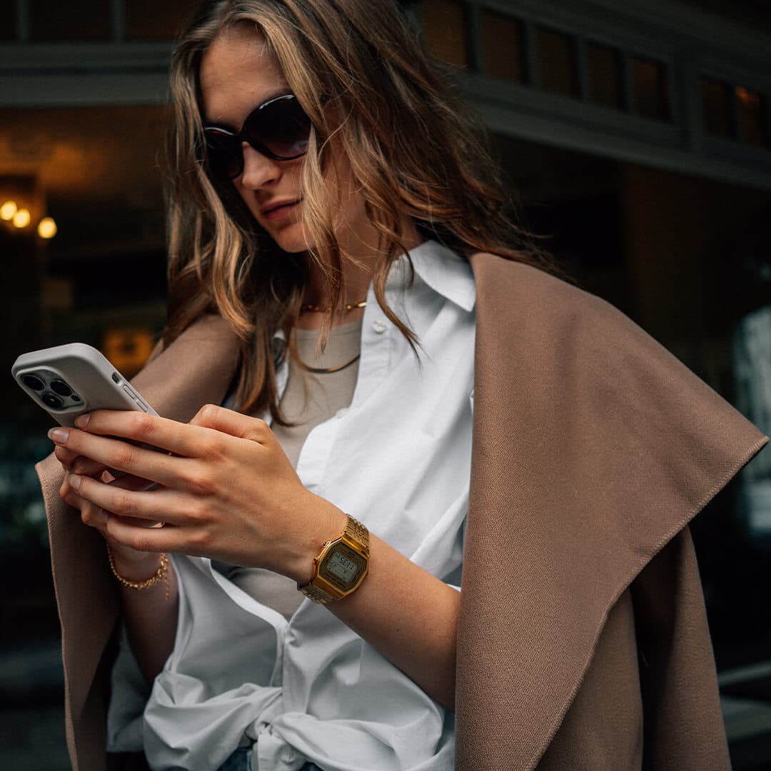Kvinne med solbriller sjekker telefonen sin, har på seg en hvit skjorte, brun frakk drapert over skuldrene og en gullklokke.