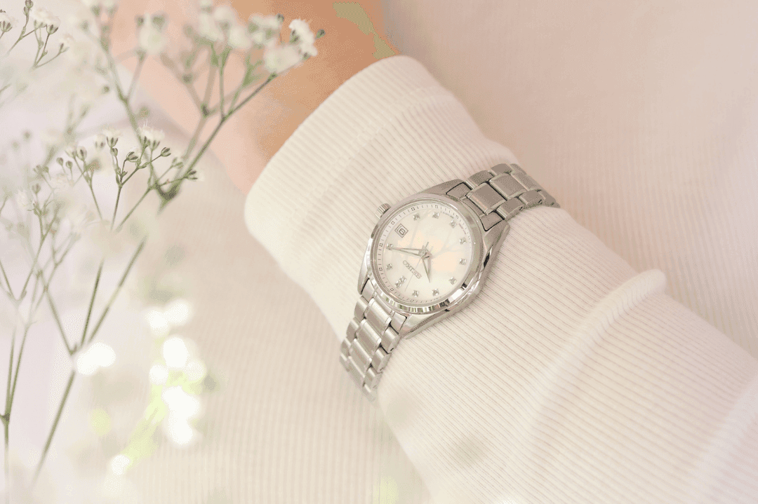 Sølvarmbåndsur med metalllenke på en persons håndledd, omgitt av delikate hvite blomster mot en myk, lys bakgrunn.