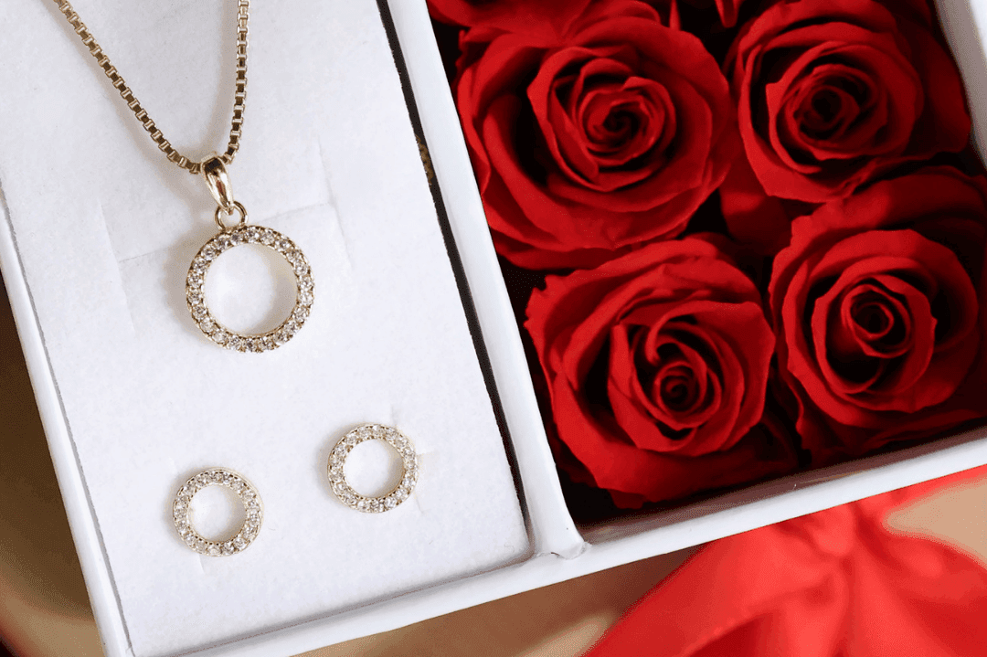 Gullkjede og øredobber med sirkulære design i en eske ved siden av livlige røde roser.