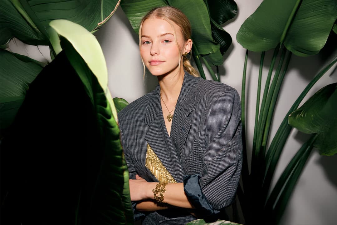 Kvinne i grå blazer med en gyllen topp under, som sitter blant store grønne blader og ser inn i kameraet med et nøytralt ansiktsuttrykk.