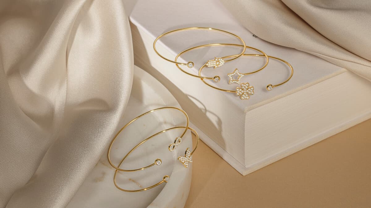 Gullarmringer med ulike anheng på en hvit bok, drapert med glatt, beige stoff, som skaper en luksuriøs og elegant ramme.