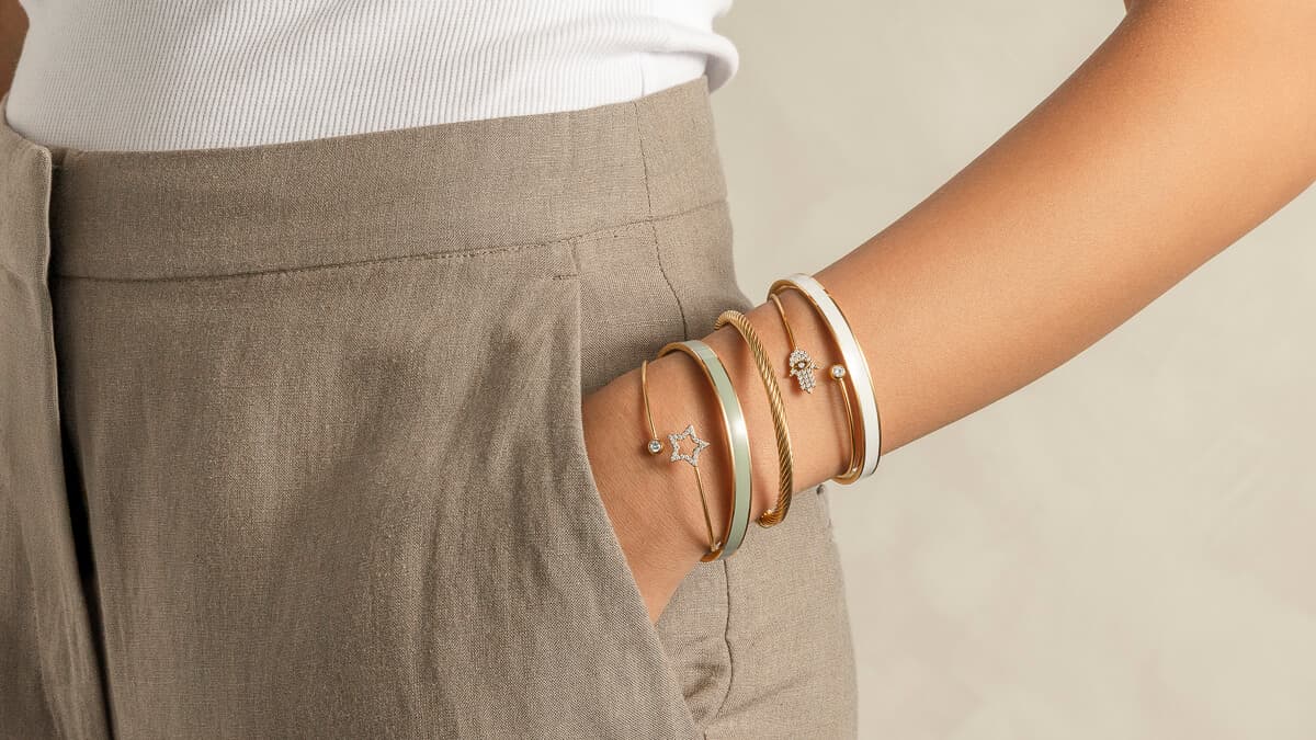 En person i beige bukser og hvit topp har hånden i lommen, og viser frem tre gullarmringer med stjerne- og perleanheng på håndleddet.
