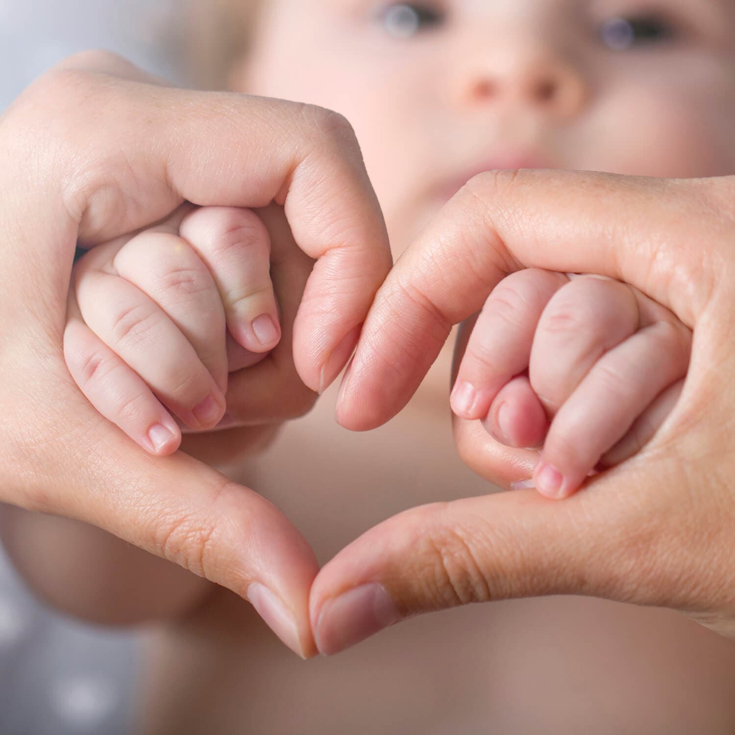 Voksen- og babyhender som former et hjerte, med babyens ansikt mykt uskarpt i bakgrunnen.
