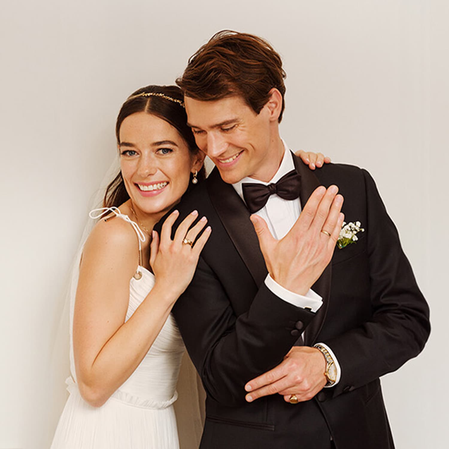 Smilende par i brudekjole, kvinne i hvit kjole lener seg på mann i svart dress. Begge viser frem ringene sine.