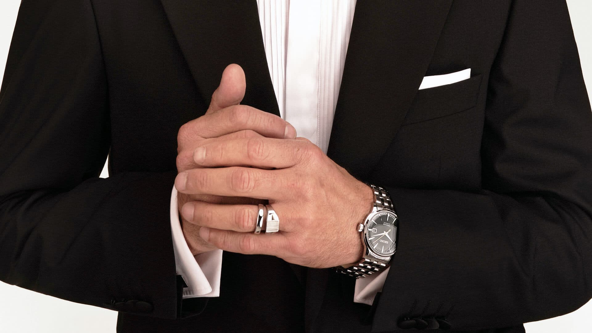 Mann i svart dress og hvit skjorte, med foldede hender. Han har på seg klokke og ring, med et synlig lommetørkle.