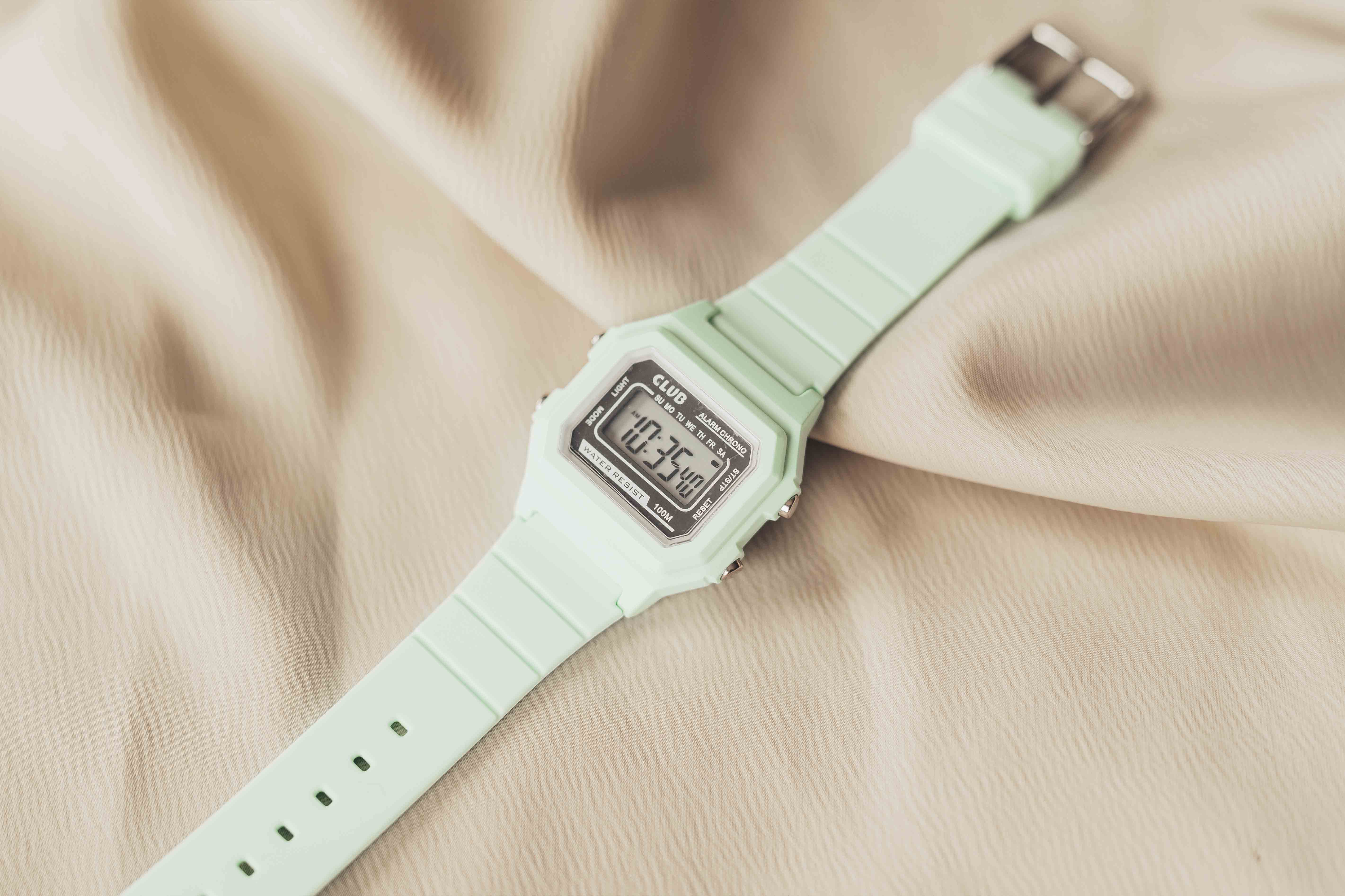 Mintgrønn digitalklokke på beige stoff, viser tiden som 10:30. Rektangulær skjerm med svarte og sølvfargede detaljer.