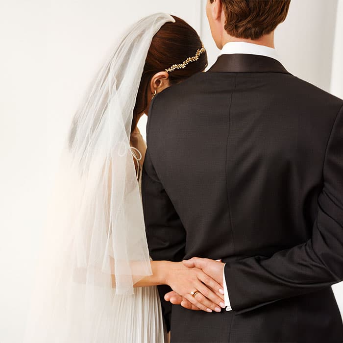 Brudeparet omfavner hverandre; bruden hviler hodet på brudgommens skulder. Hun har på slør, og han bærer svart dress.