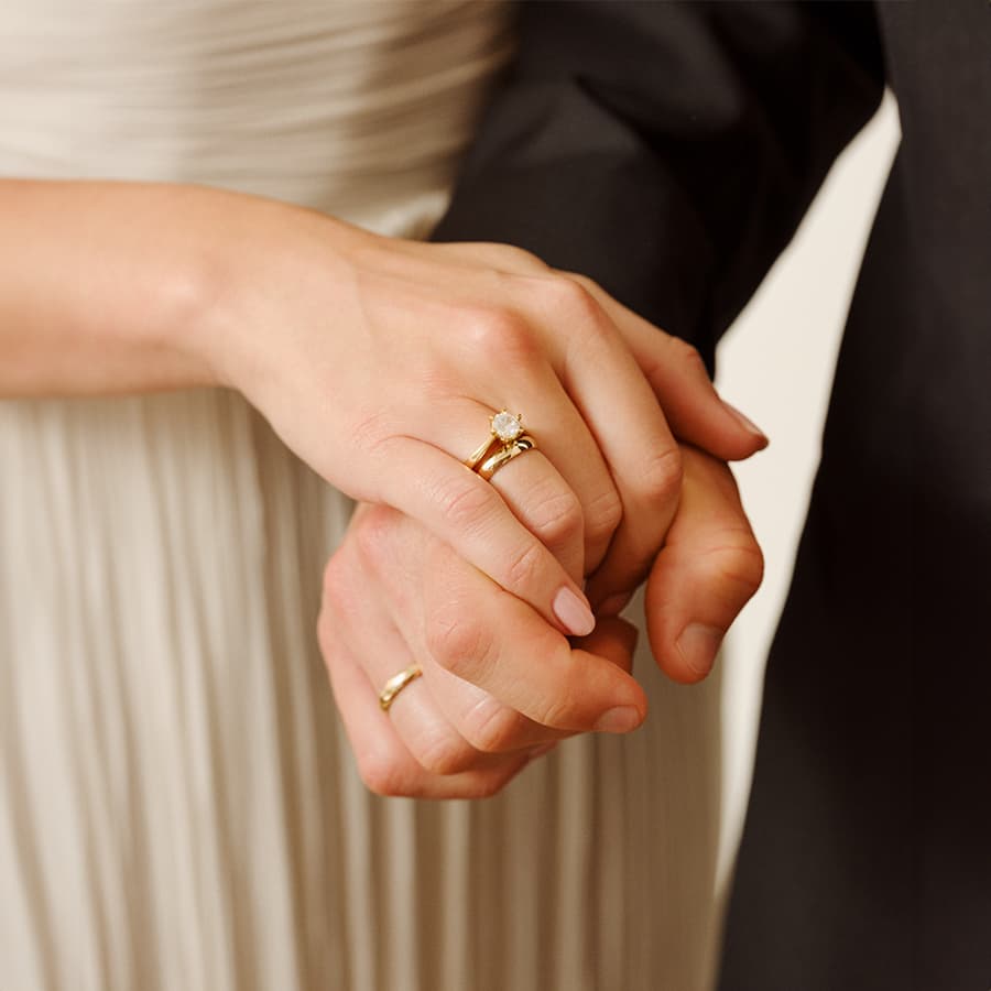 Et par som holder hender, som viser et nærbilde av gifteringene deres. Kvinnen har på seg en diamantring, og begge er elegant kledd.