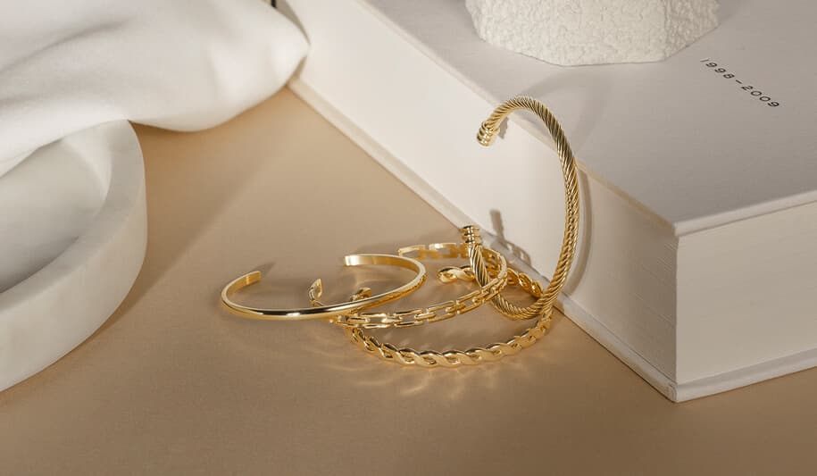 Gullarmbånd elegant arrangert på en beige overflate ved siden av en bok og mykt stoff, som kaster subtile skygger.